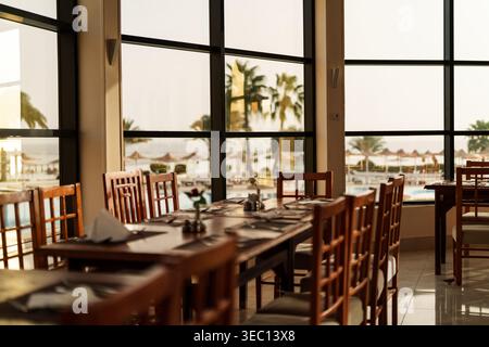 Marsa alam, egypte 21 avril 2026 : intérieur du restaurant avec tables et chaises en bois donnant sur la piscine, les palmiers et la plage sereine de la mer rouge au coucher du soleil Banque D'Images