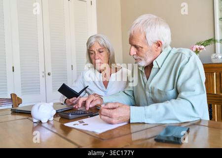 Planification de la retraite et protection financière — un couple de personnes âgées se concentre sur le calcul de ses dépenses à la maison, en utilisant une calculatrice et en comptant les pièces de monnaie Banque D'Images