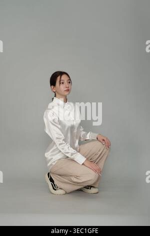 Une jeune femme dans une blouse blanche satinée et un pantalon beige s'accroupit soigneusement sur un fond neutre. Cette image souligne l'élégance de Banque D'Images