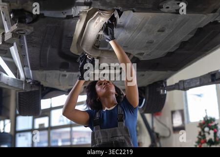 Jeune mécanicienne afro-américaine utilisant une lampe de poche pour inspecter le dessous d'une voiture soulevée sur un ascenseur dans un atelier de réparation automobile. Banque D'Images