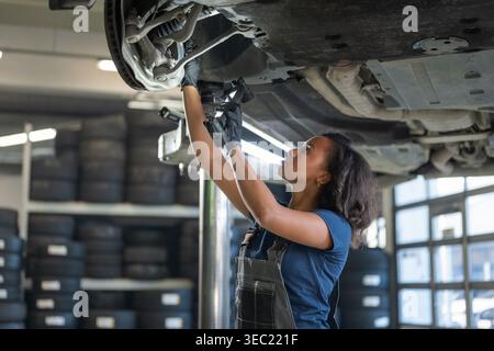 Mécanicienne concentrée utilisant une torche pour la visibilité, inspectant le dessous du véhicule dans un atelier automobile professionnel. Banque D'Images
