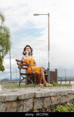 Femme dans une tenue jaune assise sur un banc en bois près du front de mer, avec ciel nuageux et montagnes en arrière-plan, profitant d'un calme et paisible Banque D'Images