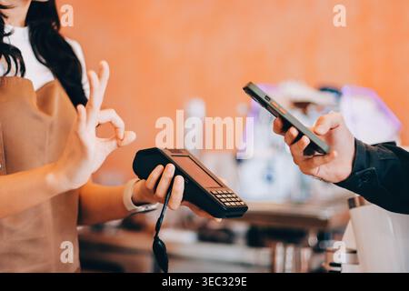 Barista adulte senior asiatique recevant un paiement mobile de la part d'un client utilisant un smartphone dans un café démontrant une transaction sans espèces moderne dans Cozy lo Banque D'Images
