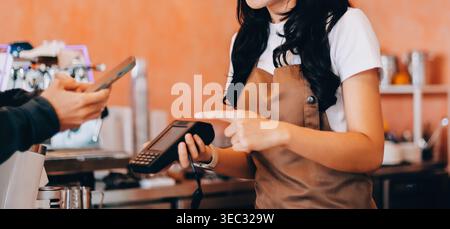 Barista adulte senior asiatique recevant un paiement mobile de la part d'un client utilisant un smartphone dans un café démontrant une transaction sans espèces moderne dans Cozy lo Banque D'Images