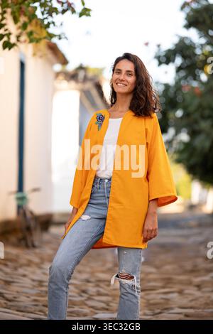 Une femme se tient debout dans une rue pavée portant un vêtement d'extérieur orange sur une chemise blanche et un Jean. Elle sourit alors qu'elle est entourée d'arbres et de bâtiments du Banque D'Images