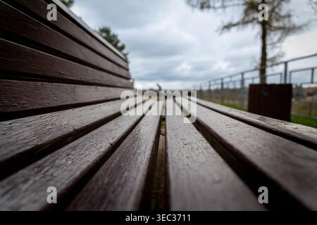 Banc en bois humide dans le parc, vue rapprochée Lowangle le long des planches trempées, gouttes de pluie sur les lattes, ciel couvert, chemin flou et garde-corps en arrière-plan, Banque D'Images