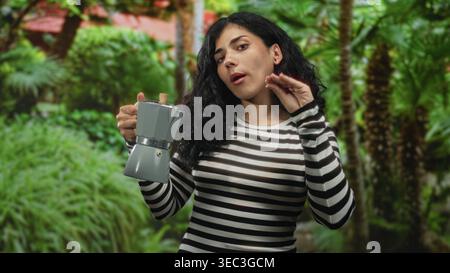 Femme tenant un pot de moka, inclinant la cafetière d'une main tout en soulevant l'autre dans un cadre forestier, haut rayé visible et les yeux fermés ; calme Banque D'Images