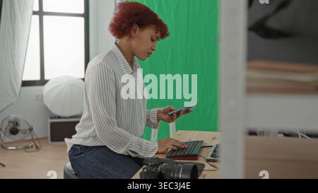 Femme tenant un smartphone et tapant sur le clavier en studio avec écran vert et caméra sur la table ; concentration créative. Banque D'Images