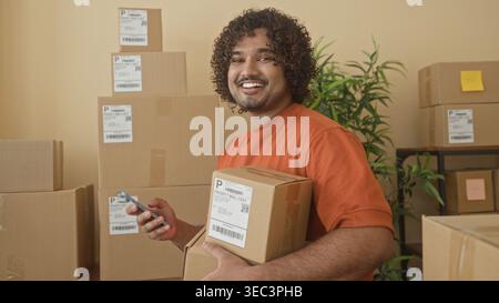 Homme tenant une boîte en carton avec smartphone à la main, souriant au milieu de colis empilés, étiquettes d'expédition et une plante en pot sur les étagères à l'intérieur d'un dépôt de bâtiment Banque D'Images