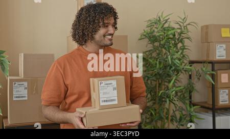 L'homme tient deux paquets de carton avec les mains, souriant et montrant l'étiquette d'expédition sur la boîte supérieure dans une zone de parcelle de bâtiment avec des boîtes empilées et une étagère Banque D'Images