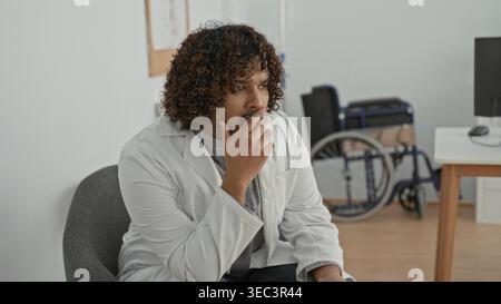 Homme médecin en manteau blanc assis, tenant le stéthoscope dans les mains, geste de toux de la main à la bouche, fauteuil roulant visible près du bureau dans le bâtiment de la clinique ; concer Banque D'Images