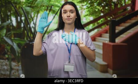 Femme infirmière en gommage portant des gants bleus lève la main droite et place la main gauche sur la poitrine dans un bâtiment ; devoir. Banque D'Images