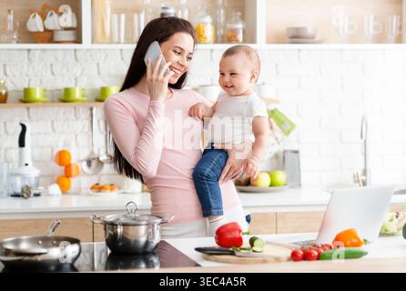 Bonne mère parlant au téléphone tout en tenant bébé dans la cuisine moderne préparer le repas Banque D'Images