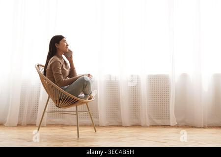 Femme réfléchie assise dans une chaise moderne par une grande fenêtre avec des rideaux transparents Banque D'Images