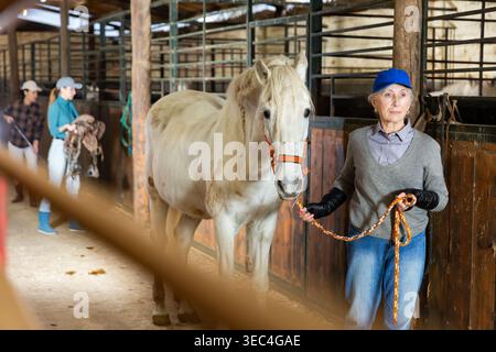 Ferme femelle âgée tenant des reins menant le cheval blanc à travers stable Banque D'Images