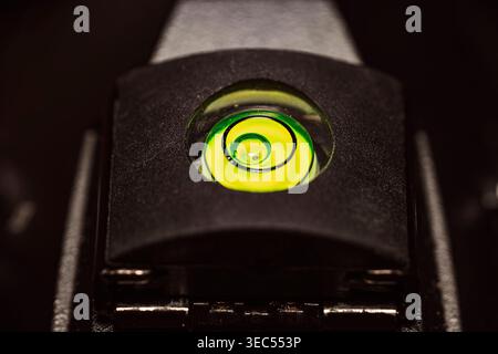 Photographie macro extrême mettant en évidence un niveau à bulle circulaire vert et jaune vif monté sur une tête de trépied noire dans un environnement de faible luminosité Banque D'Images