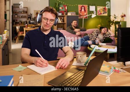 Jeune homme caucasien adulte étudiant avec des écouteurs, écrivant des notes à côté d'un ordinateur portable tandis que des colocataires se détendent derrière, montrant la concentration à la maison. Utile pour la vie étudiante, la préparation aux examens, le concept de distraction Banque D'Images
