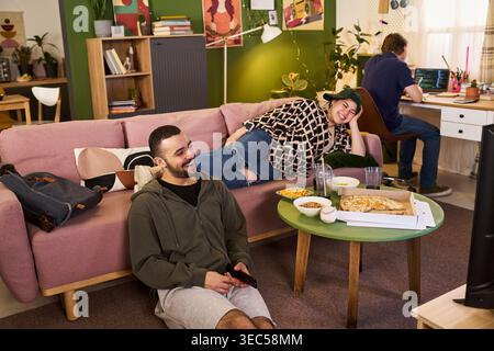 Jeunes colocataires adultes se détendant avec pizza et jeu vidéo tandis que l'étudiant masculin code sur ordinateur portable en arrière-plan, montrant un logement étudiant partagé. Idéal pour le style de vie du campus, la vie des jeunes, le travail d'équipe Banque D'Images