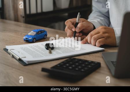 Homme signant le document de police d'assurance automobile avec stylo à côté de la clé de voiture, véhicule modèle, calculatrice et ordinateur portable sur le bureau, représentant l'approbation de la police, couverture Banque D'Images