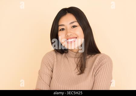 Sourire jeune femme avec de longs cheveux noirs portant un pull beige sur un fond clair Banque D'Images