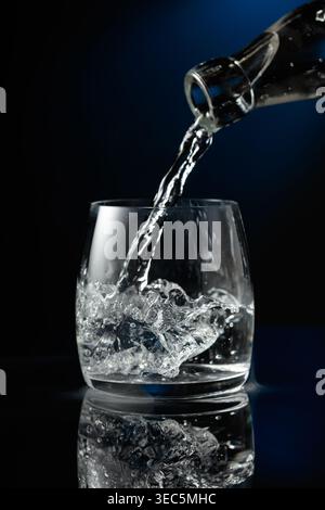 Vodka ou gin limpide versé sur de la glace dans un verre. Vodka avec de la glace sur un fond réfléchissant noir. Banque D'Images