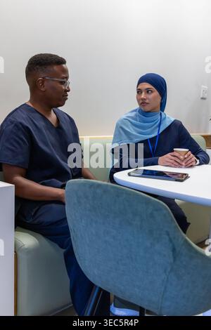 Divers collègues portant des gommages bleu marine et du hijab assis dans la salle du personnel à l'aide d'une tablette et d'un gobelet de maintien Banque D'Images