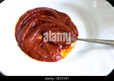 La sauce rouge tourbillonne dans un bol blanc. Cuillère en métal repose à côté de la pâte épaisse. La sauce scintille sous un éclairage doux. Bol a des bords subtils et courbés. Surface Banque D'Images