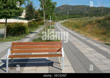 Viladecans, Espagne - 29 avril 2026 : un banc de parc moderne en bois se trouve le long d'un sentier piétonnier pavé sinueux dans un paysage méditerranéen mettant en vedette des jeunes Banque D'Images