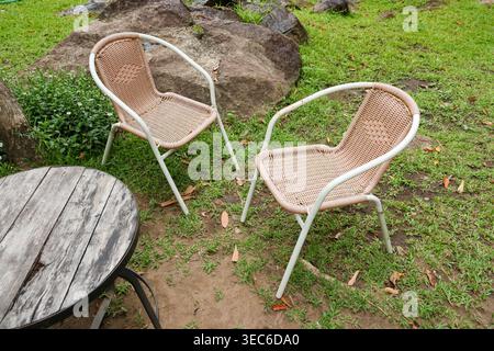 Cadre paisible de jardin extérieur avec chaise tissée vide à côté de la table rustique en bois sur l'herbe verte créant une atmosphère sereine relaxante pour c calme Banque D'Images