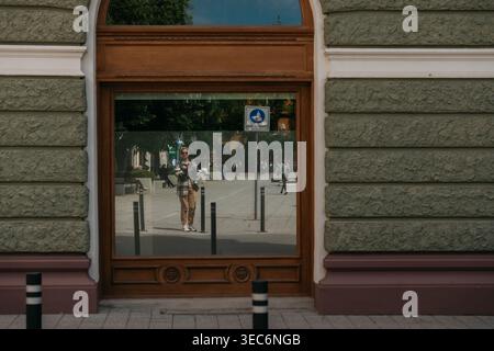 Femme prenant un selfie reflété dans une grande fenêtre de rue tandis que les piétons marchent à travers le centre de Subotica. Subotica, Serbie - 27 avril 2026. Banque D'Images