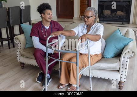 Femme afro-américaine senior assise sur le canapé à la maison à l'aide d'un marcheur, soignant dans l'observation des gommages Banque D'Images