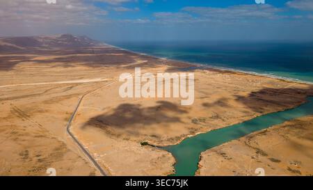 Vue aérienne de la côte désertique aride avec un lagon turquoise sinueux ou une rivière rencontrant l'océan bleu profond sous des nuages éparpillés. Banque D'Images