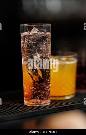 Deux cocktails rafraîchissants de longue boisson remplis de glaçons et de liquide gazéifié sont assis sur un comptoir de bar noir texturé. Banque D'Images