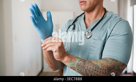 Tatoué Doctor mettant sur des gants médicaux bleus avec stéthoscope dans un cadre clinique Banque D'Images