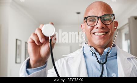 Médecin masculin souriant avec stéthoscope tenant l'instrument vers la caméra dans la clinique à domicile Banque D'Images