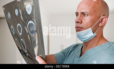 Médecin homme inquiet examinant le film d'IRM cérébrale en clinique portant un masque chirurgical et des gommages Banque D'Images