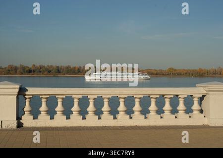 Un bateau de croisière fluviale de passagers navigue le long d'une rivière calme en automne, vu d'une promenade en bord de mer avec une balustrade blanche, un ciel bleu doux. Banque D'Images