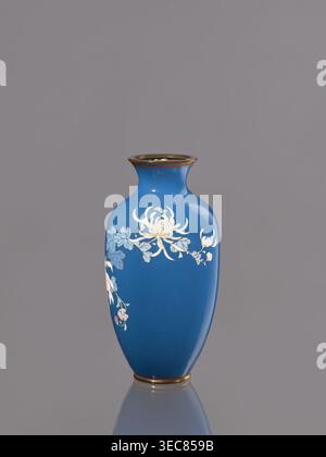 Vase avec chrysanthèmes, CA. 1900 Meiji 23, Japon, Nagoya ; récipient en émail cloisonné ; hauteur 18,0 cm, diamètre 9,0 cm., Un vase bleu avec un col étroit et un bord évasé repose sur un fond gris, montrant une surface brillante et une base réfléchissante. Le vase présente un motif floral avec des fleurs jaunes pâles et blanches et des feuilles bleu-vert, et de petites touches de rose sur certains bourgeons. La jante et la base ont une bande d'aspect métallique dans un ton doré brunâtre chaud. Banque D'Images
