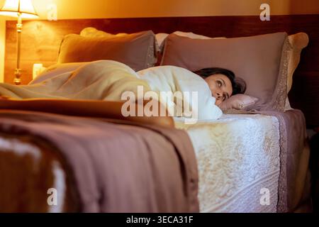 Le matin, une jeune femme portant une robe est couchée dans le lit. Elle regarde par la fenêtre, regardant le monde extérieur. La chambre dispose d'une lumière chaude et confortable b Banque D'Images