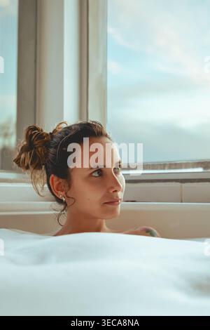 Une jeune femme aux cheveux attachés est assise dans un bain moussant, regardant par la fenêtre. Une lumière douce remplit la pièce, créant une atmosphère simple. Elle aime un quie Banque D'Images