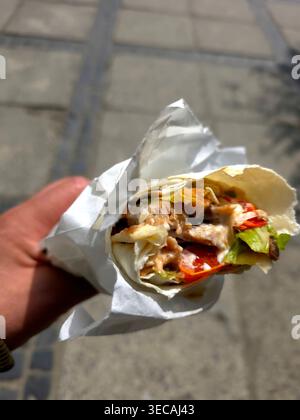 Une personne tient un kebab enveloppé dans sa main tout en se tenant debout sur le trottoir. Le kebab comprend de la viande, de la laitue et de la sauce. Banque D'Images