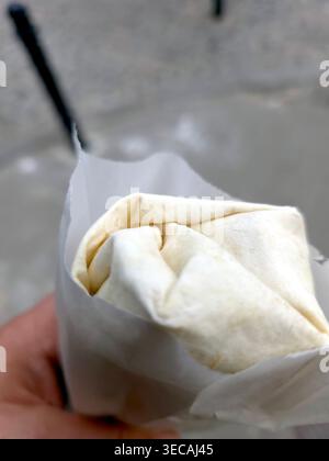Une main tient un wrap rempli de kebab et de shawarma pendant que les gens passent dans une rue animée dans l'après-midi. La scène montre une atmosphère animée. Banque D'Images
