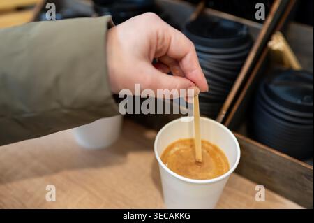 Gros plan d'une main féminine en veste verte mélangeant un cappuccino ou un latte frais avec un agitateur jetable dans un gobelet à emporter dans une station libre-service de café Banque D'Images
