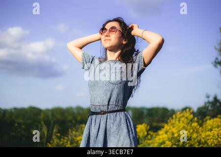 Une femme se tient dans un champ rempli de fleurs jaunes portant une robe en denim et des lunettes de soleil elle lève les bras et regarde le ciel sur un ensoleillé clair Banque D'Images