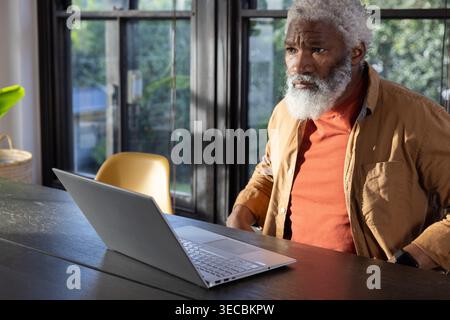 Homme afro-américain senior assis à la maison à l'aide d'un ordinateur portable par les fenêtres dans la chemise de rouille, espace de copie Banque D'Images