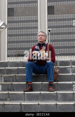 Homme d'âge moyen assis sur des marches en béton à l'extérieur du bureau tenant à emporter et serviette avec sac bronzé Banque D'Images