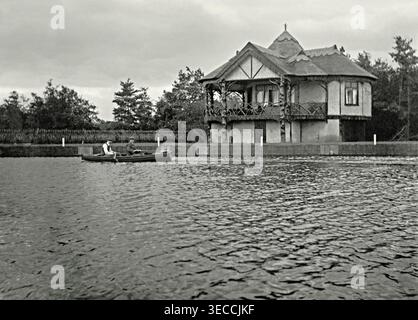 Une première photographie avec deux hommes dans un bateau à rames sur la rivière Bure en passant par « The Glade » à Wroxham, Norfolk, Angleterre, Royaume-Uni. La charmante maison de style rustique contient un hangar à bateaux et une véranda profonde. Il a été construit à l'aide de troncs d'arbres noueux et d'un treillis de branches d'arbres contorgées, avec un toit de chaume de roseaux de Norfolk. Le Glade est une maison au bord de la rivière, connue à l'origine sous le nom de « Bungalow espagnol ». Il a été construit vers 1900 pendant une période de construction de maisons de vacances récréatives et de hangars à bateaux sur des terrains vendus par le domaine Wroxham House. Banque D'Images
