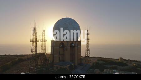 De grandes antennes radar et des tours de communication dominent le paysage d’une installation militaire côtière, silhouettée contre les teintes chaudes d’un soleil couchant sur la mer calme. Vol de drone Banque D'Images