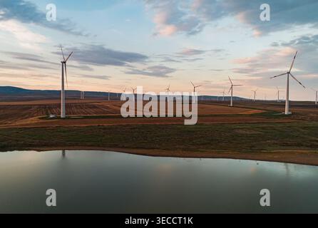 La vue aérienne d'une éolienne génère de l'énergie propre pendant le coucher du soleil sur un lac tranquille dans un paysage rural avec des champs ouverts Banque D'Images