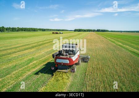 La vue aérienne capture la puissante moissonneuse-batteuse rassemble les récoltes sur un vaste champ vert sous un ciel bleu clair lors d'une journée ensoleillée à proximité Banque D'Images